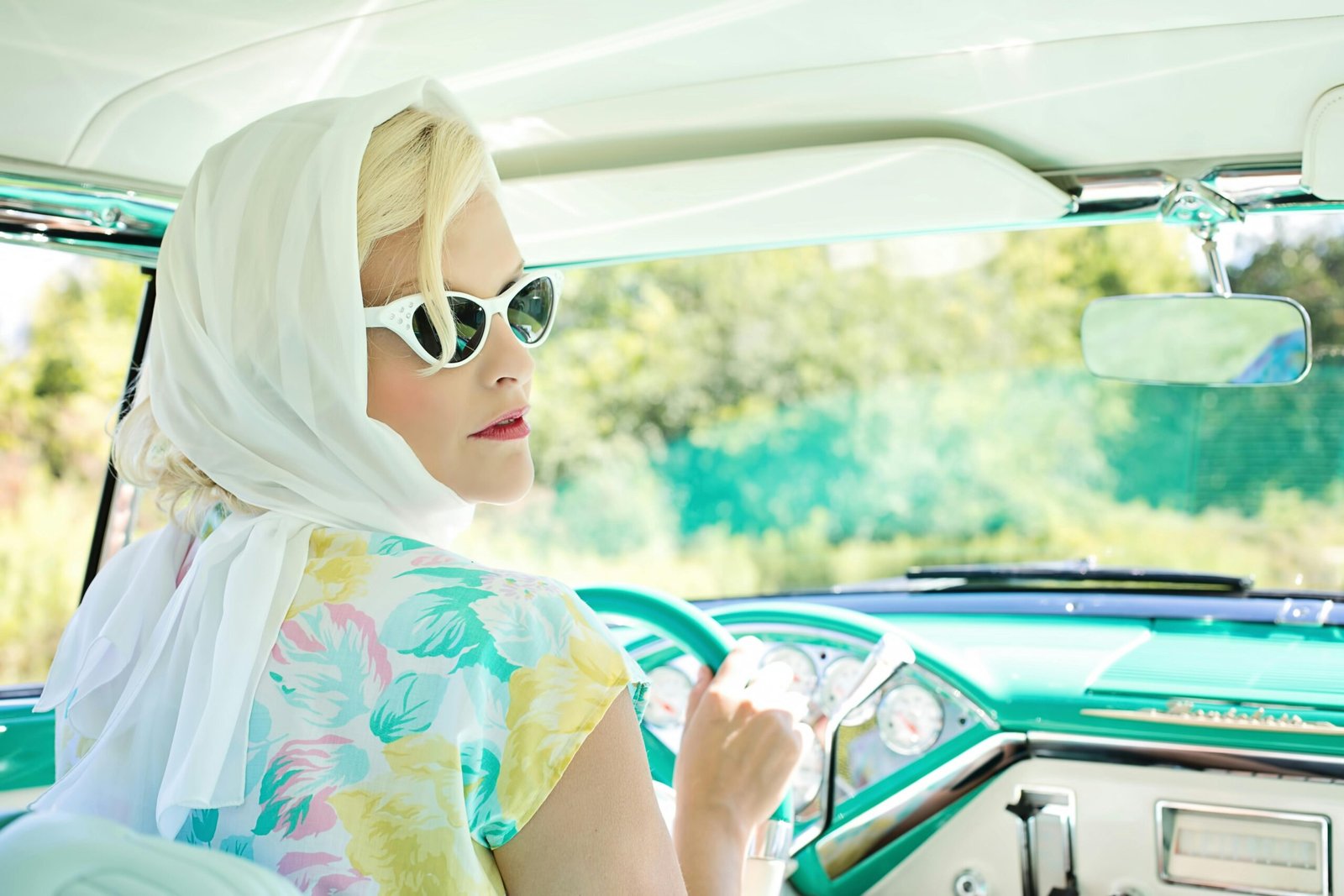 Femme élégante au style rétro dans une voiture vintage, portant foulard blanc et lunettes œil de chat.